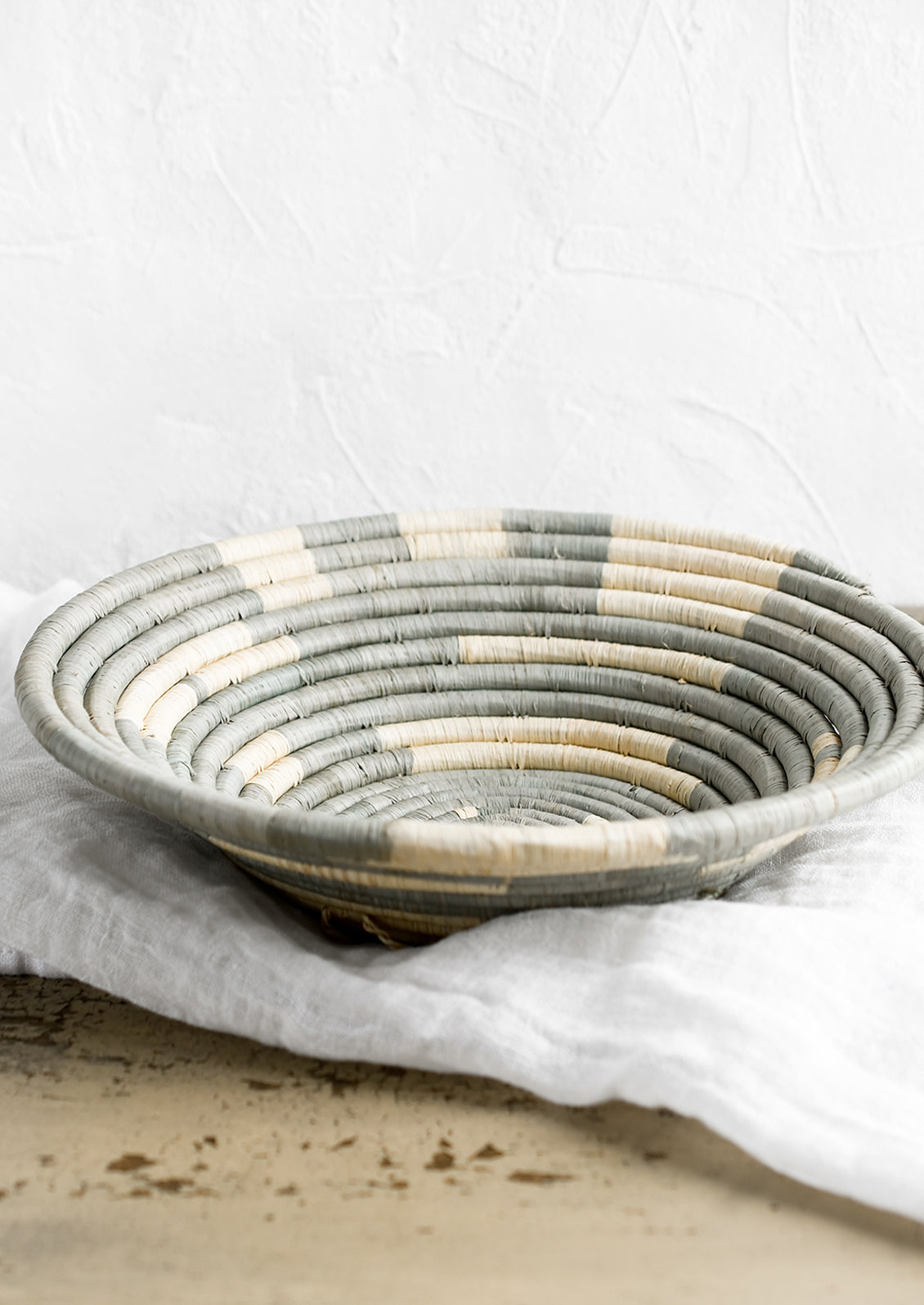 A round raffia bowl in dusty blue-grey and natural spiral geometric pattern.