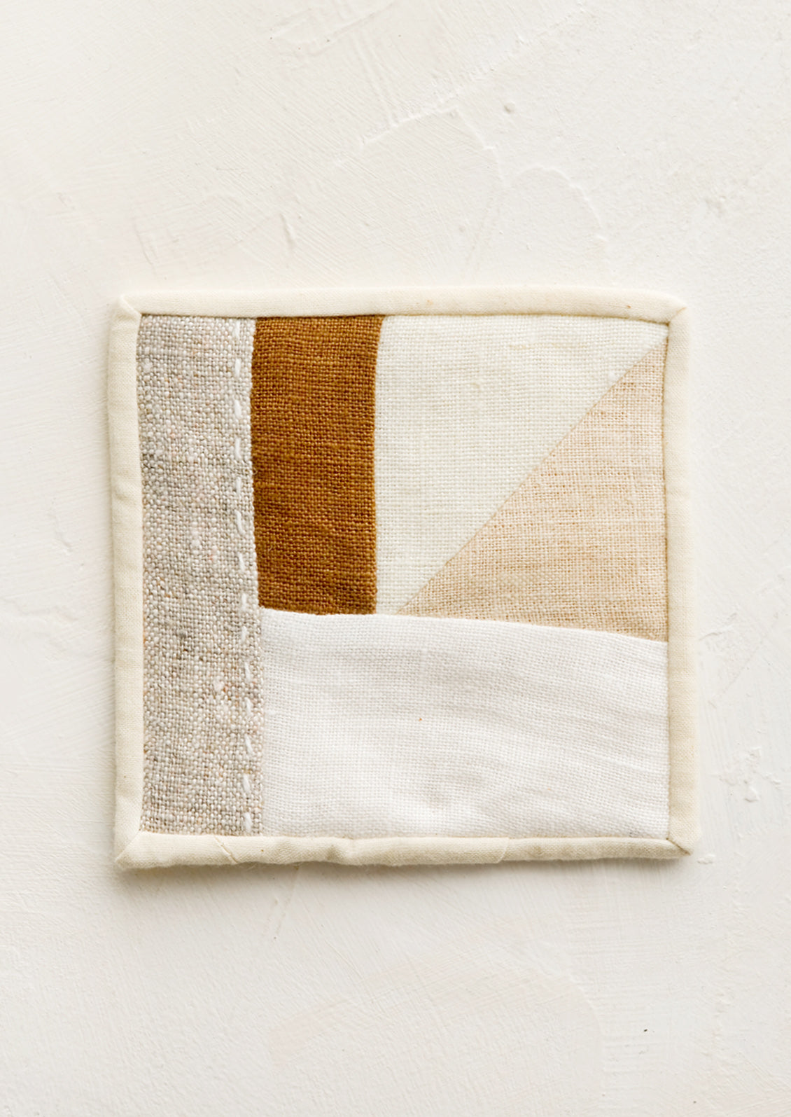 A square drink coaster made from neutral hued patchwork fabric.