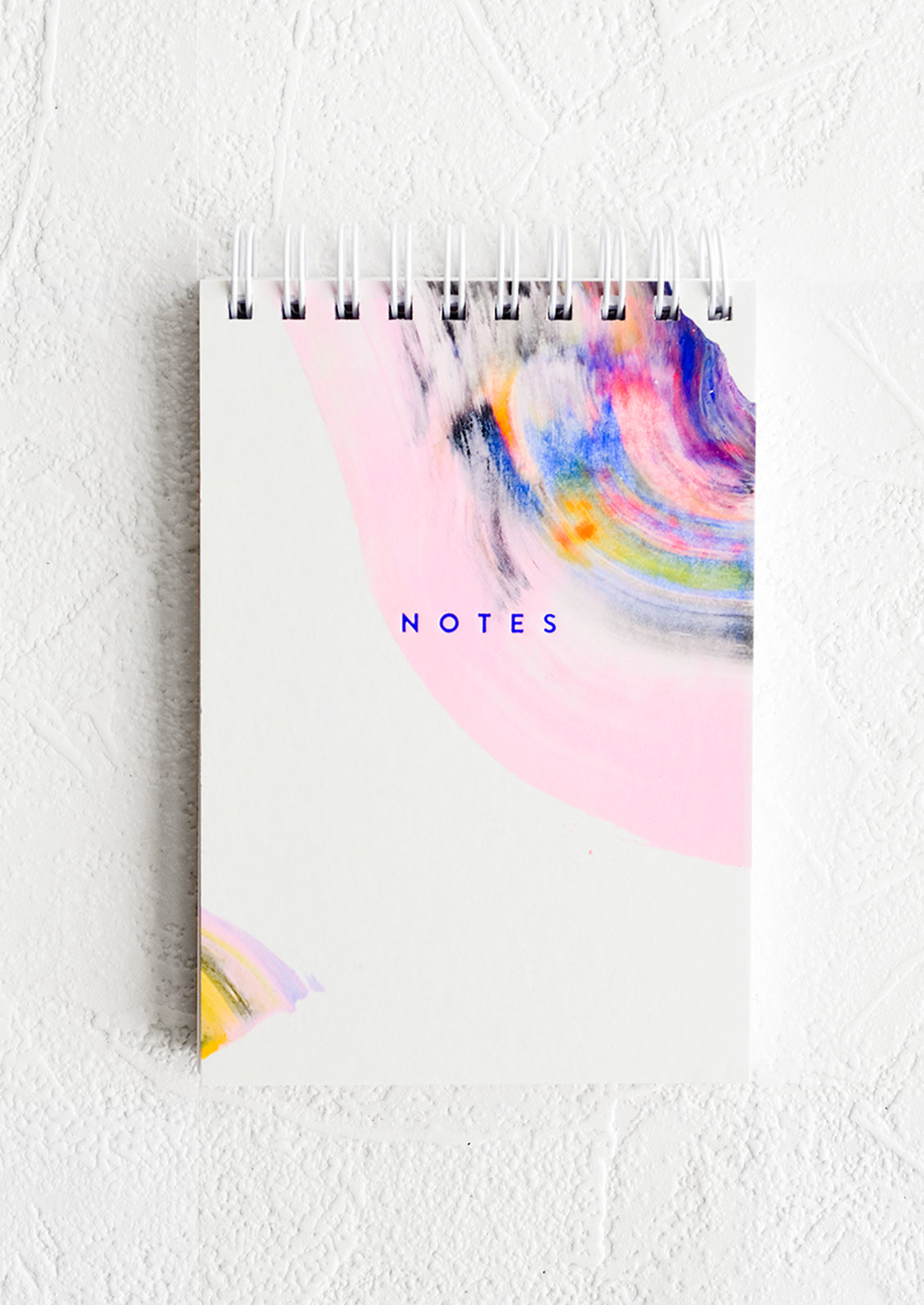A small white spiral bound notepad with rainbow swirl painted cover and "NOTES" on front.