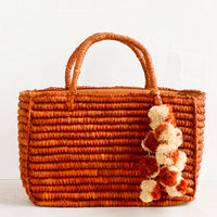 Cinnamon: An east-west raffia tote in rust color with straw pom pom detailing.
