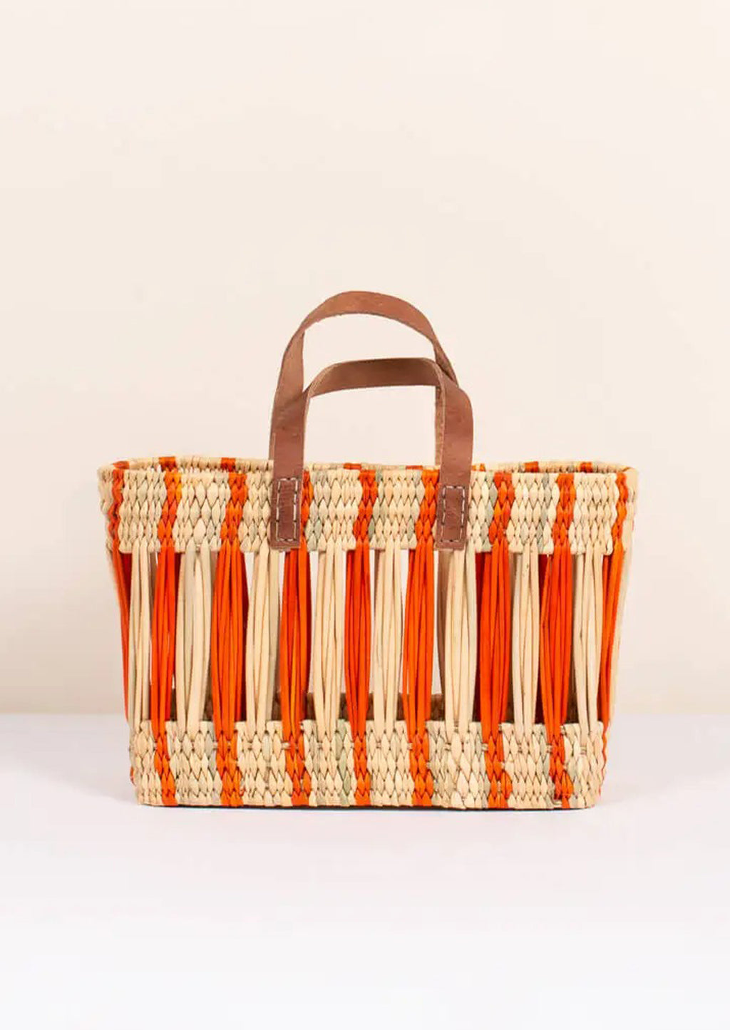 Small / Clementine: An oblong open weave basket in natural reed with bright orange stripes and brown leather handles.