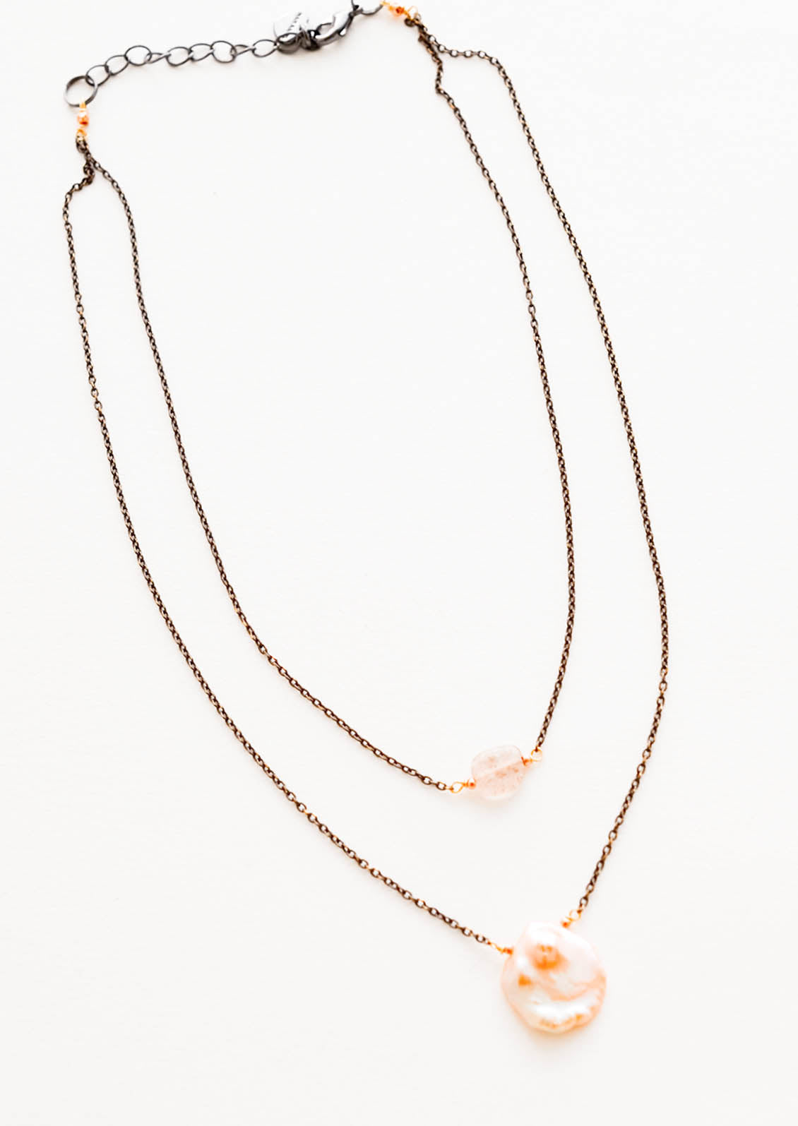 Two layer necklace composed of one dark gold chain with translucent pink crystal pendant and one of dark gold chain and pink freshwater pearl pendant.