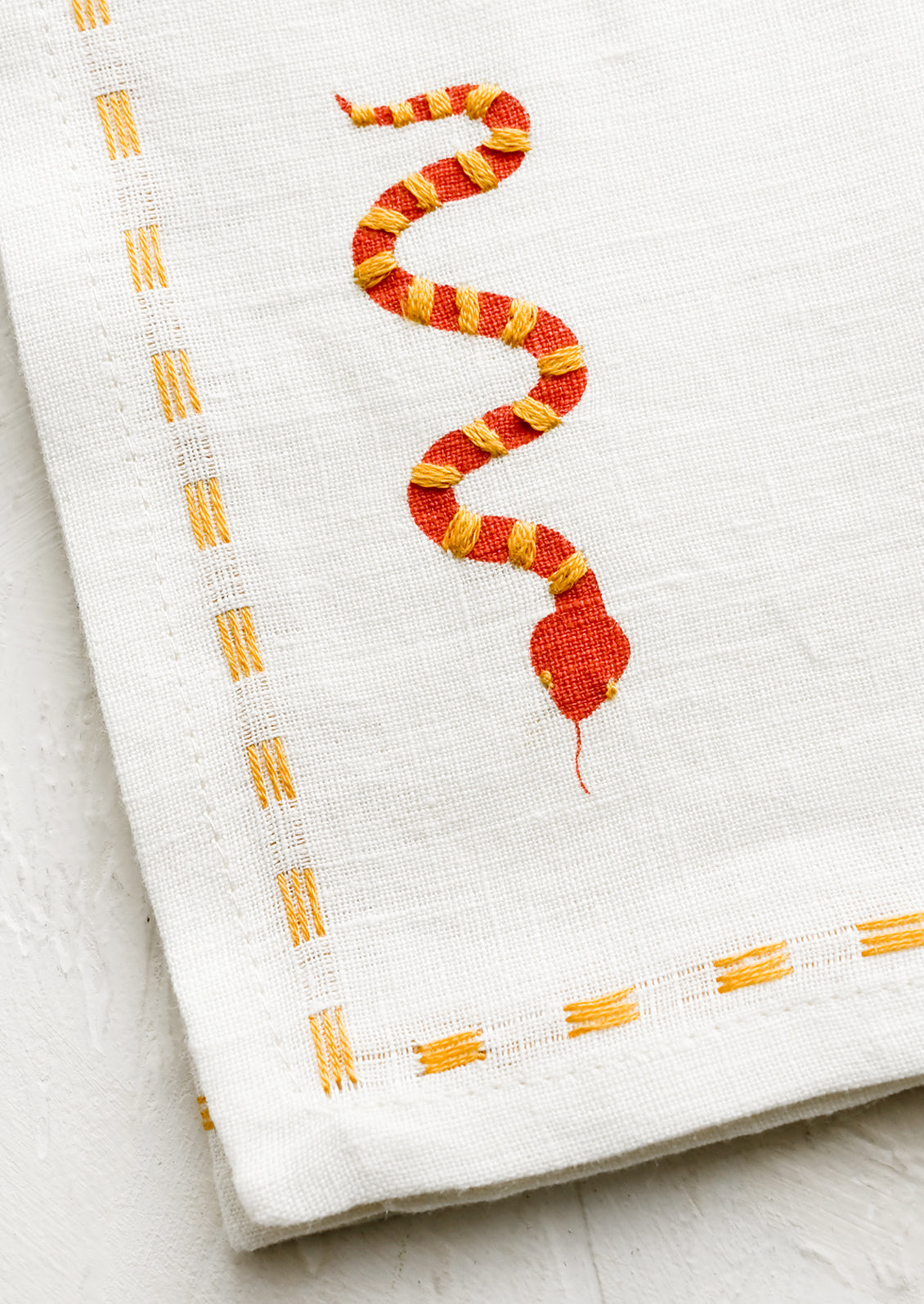 A white linen napkin with yellow stitching and red and yellow snake embroidery at corner.