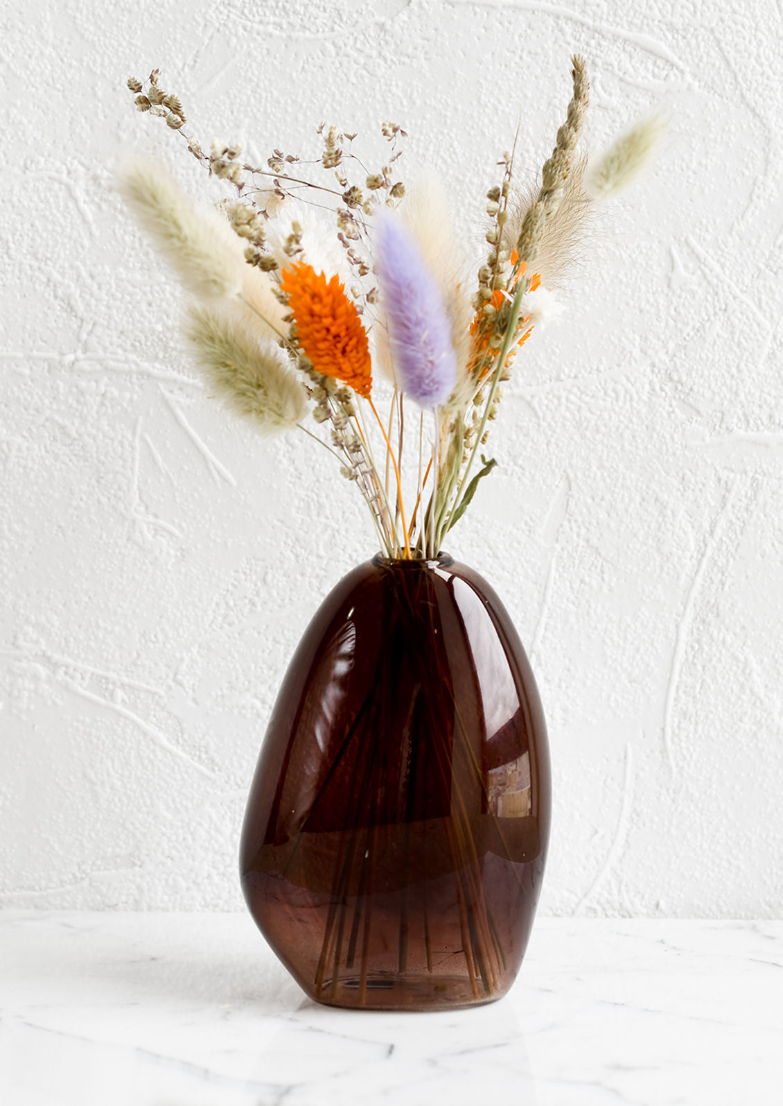 An asymmetrical bud vase in brown colored glass, holding dried flowers.