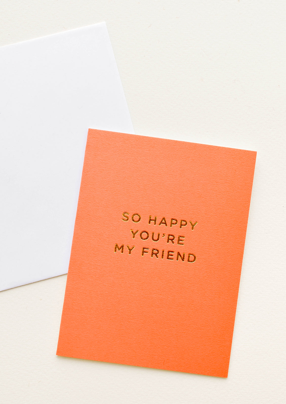 A white envelope and orange greeting card with the words "so happy you're my friend" in gold foil.