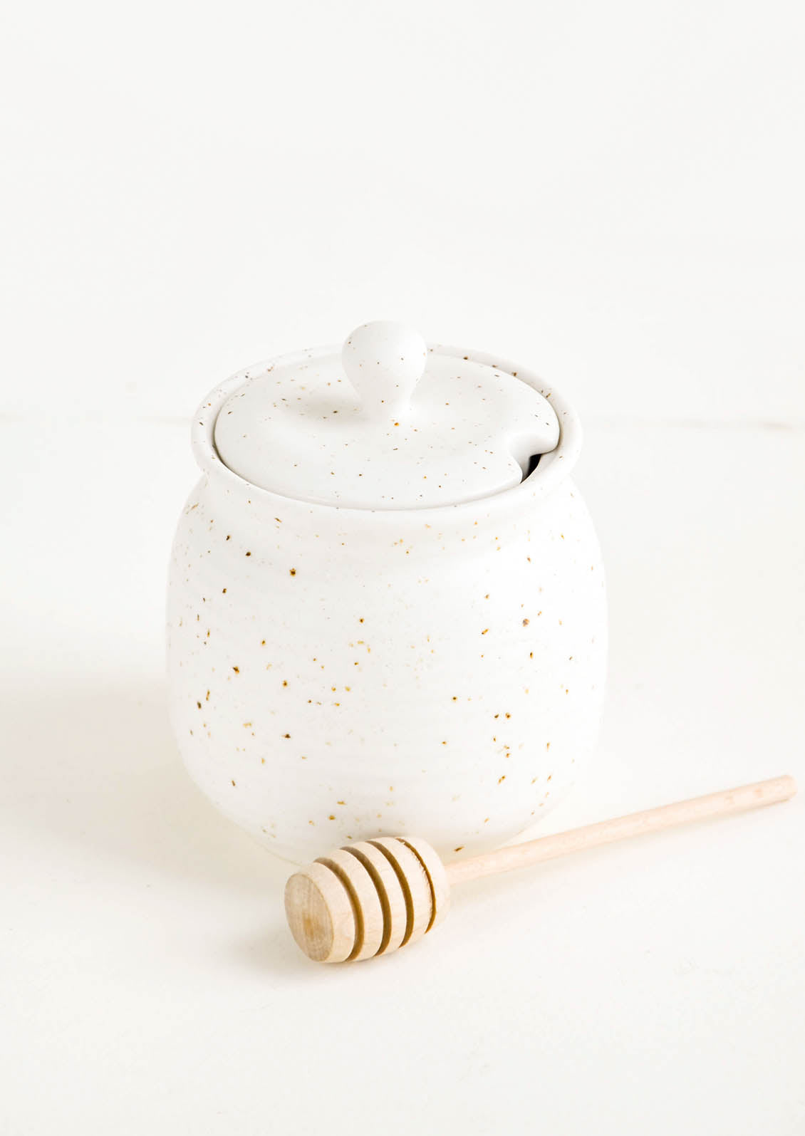 Round ceramic honey jar in speckled white glaze with lid and wooden dipper