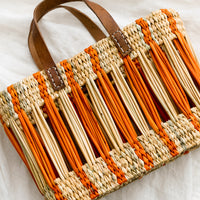 1: An oblong open weave basket in natural reed with bright orange stripes and brown leather handles.