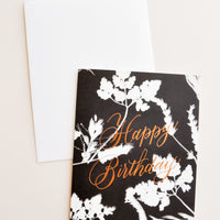 1: Notecard with white flowers on black background and the text 'Happy Birthday" in metallic script, with white envelope.