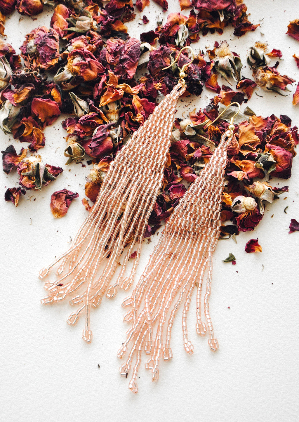 Iridescent Blush: Light pink iridescent beaded fringe earrings rest atop a bed of dried rose petals.