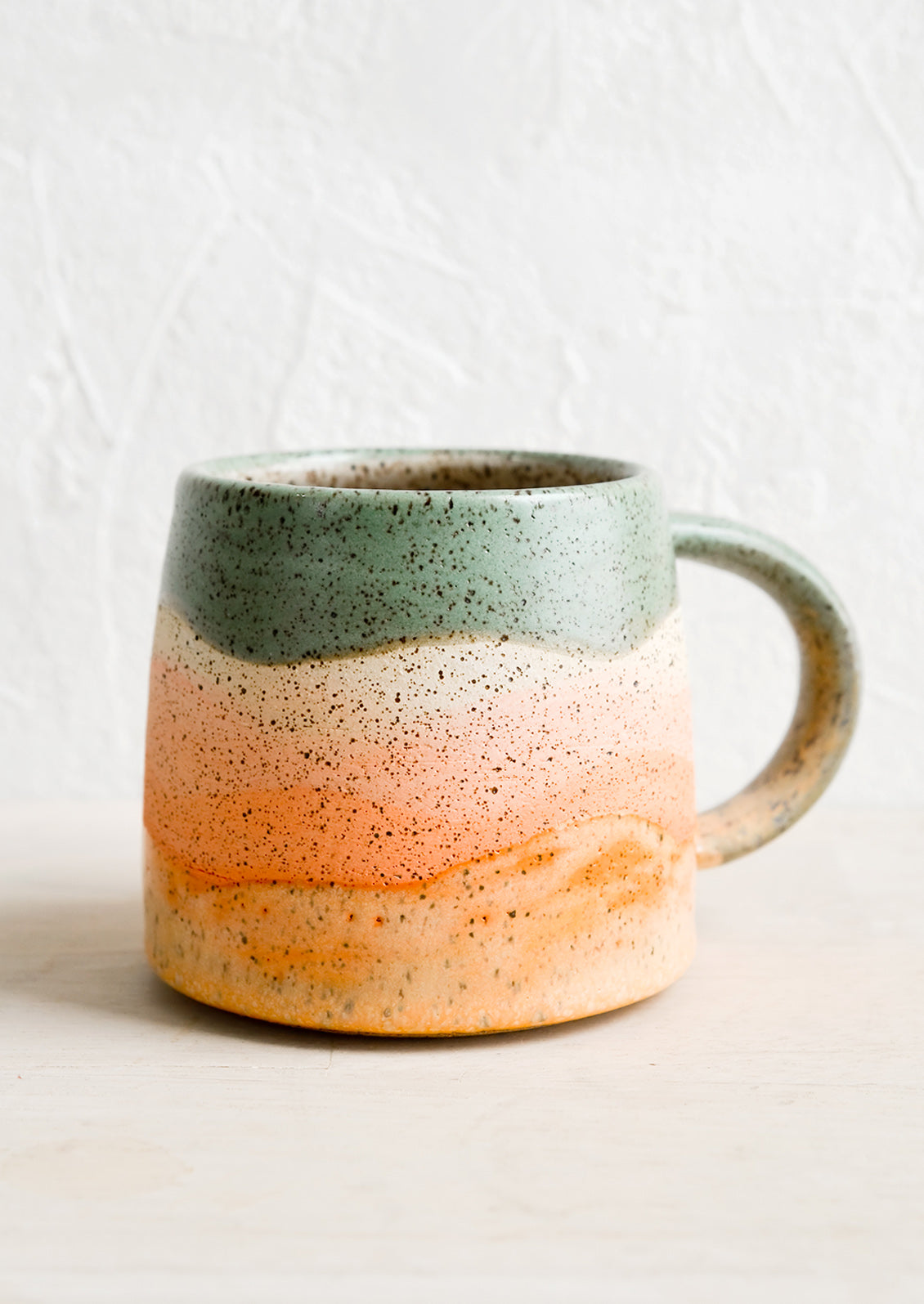A ceramic mug with handle in wavy stripe design in celadon, coral and orange.