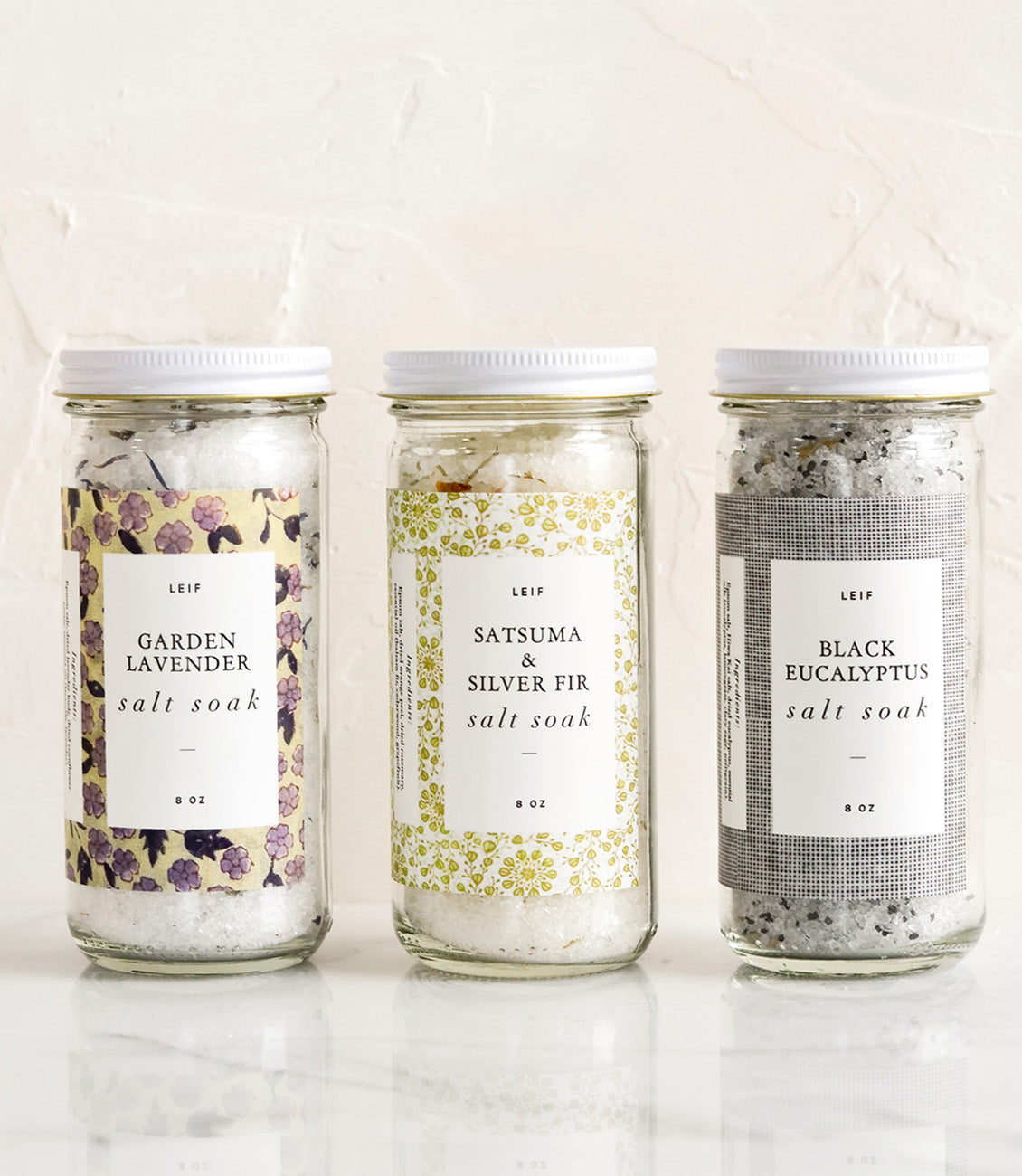 Three glass jars with decorative labels containing bath salt soaks.