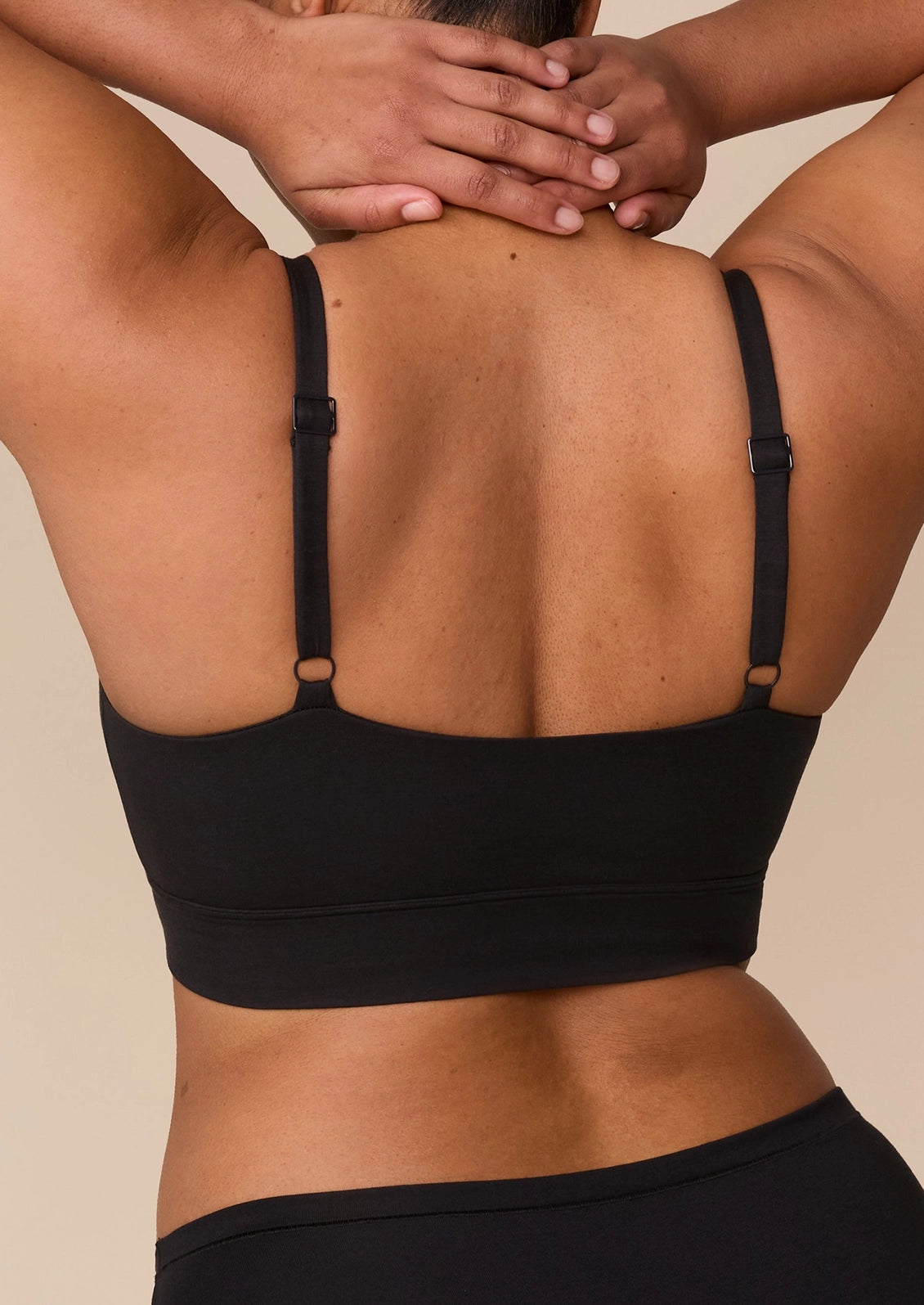 Rear view of a model wearing a black sports bra with adjustable underband straps and a wide supportive band, paired with black leggings, against a solid beige background.