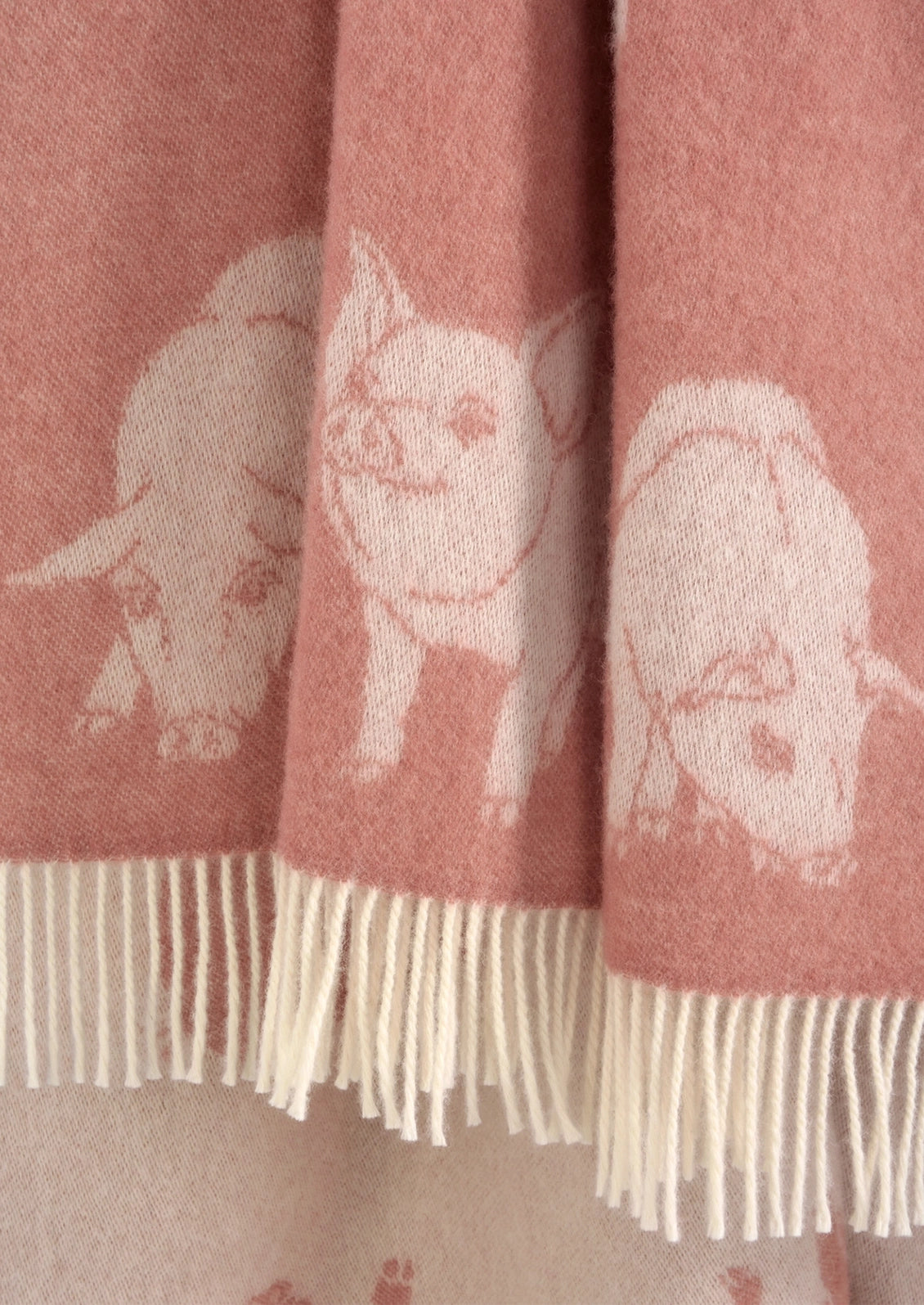 A close-up of a dusty pink knit blanket featuring cream-colored pig illustrations and matching cream fringe along the bottom edge.
