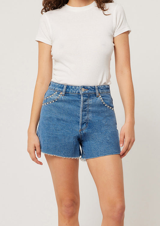 Person wearing a white t-shirt and blue denim shorts with embellishments on a plain background