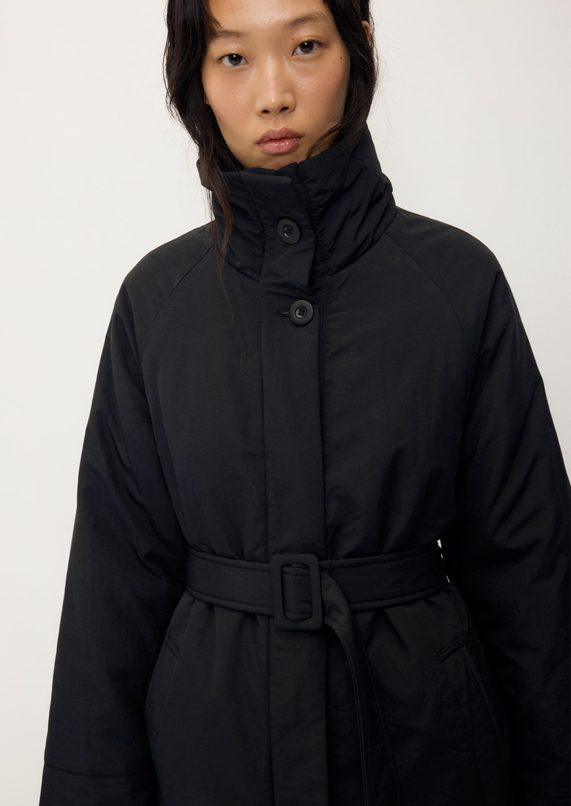 A model wearing a black high-neck belted coat with a high collar and front buttons, photographed against a plain light gray background.