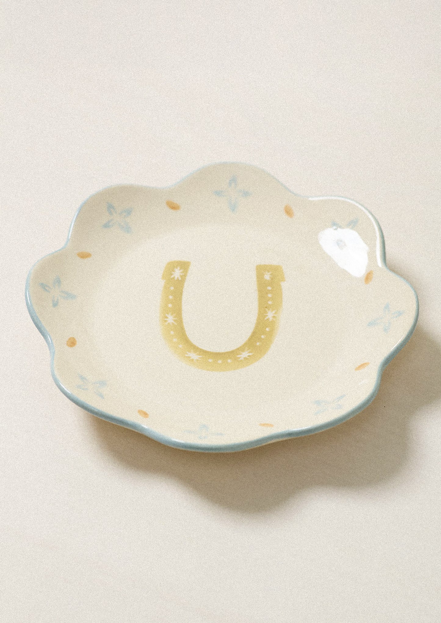 A scalloped ceramic dish with a yellow horseshoe design in the center and light blue floral accents around the rim, set against a warm beige background.