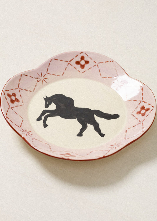 A scalloped ceramic plate with a pink and red patterned rim and a black silhouette of a rearing horse in the center, set against a light beige background.