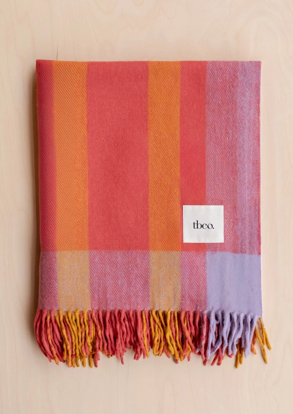 A multicolor woven throw blanket with fringe, featuring vertical stripes of coral, orange, yellow, pink, and lavender, laid flat on a light wood background. A white square label with the brand name 'thco.