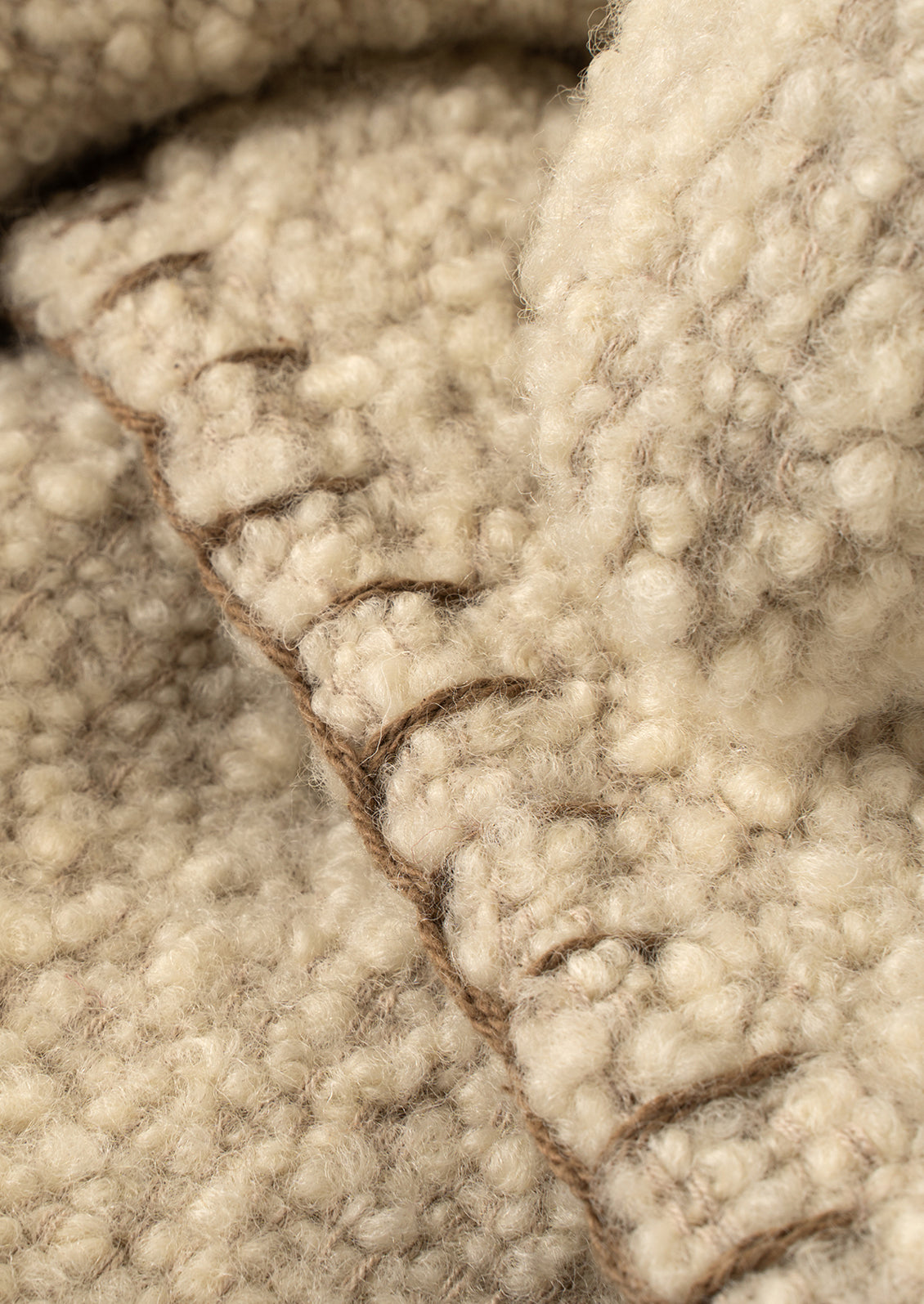 Close-up of a textured cream-colored wool fabric with a stitched seam in a warm brown tone.