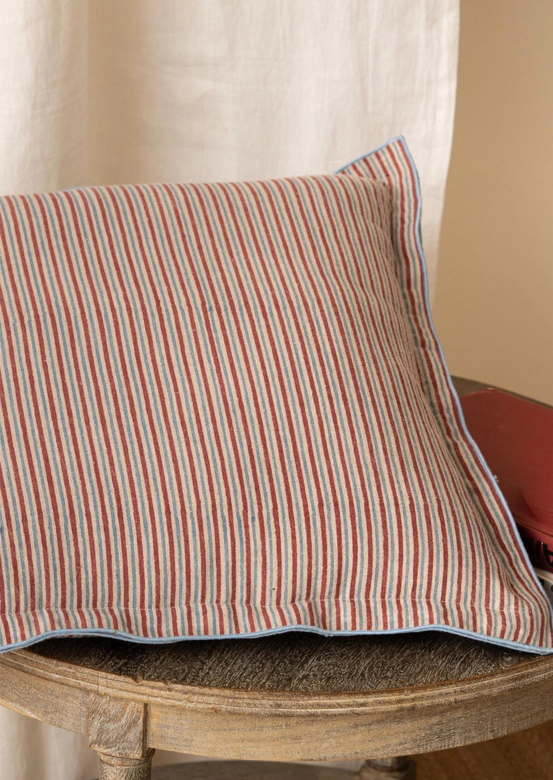 A square throw pillow with vertical red and blue stripes on a beige fabric background, finished with a solid blue piped border.