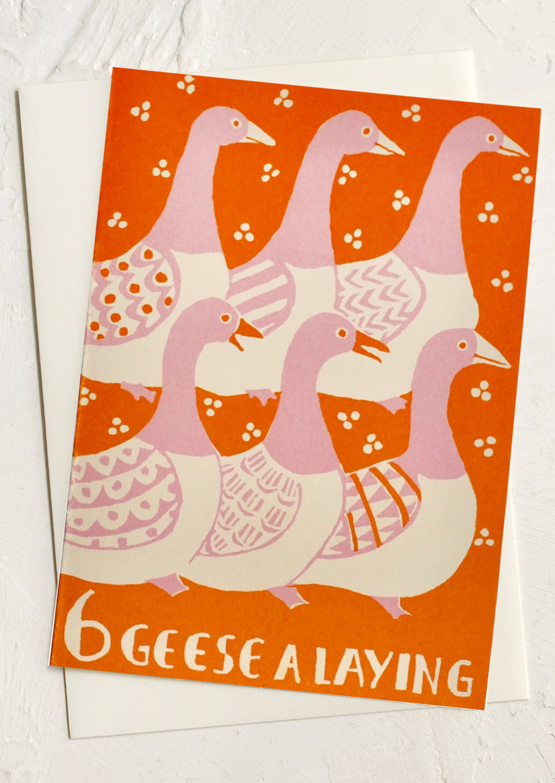 A vintage-style greeting card featuring an illustration of six pink and cream geese on an orange background, with the text '6 GEES E A LAYING' at the bottom. The card is displayed on top of a white envelope against a light textured background.
