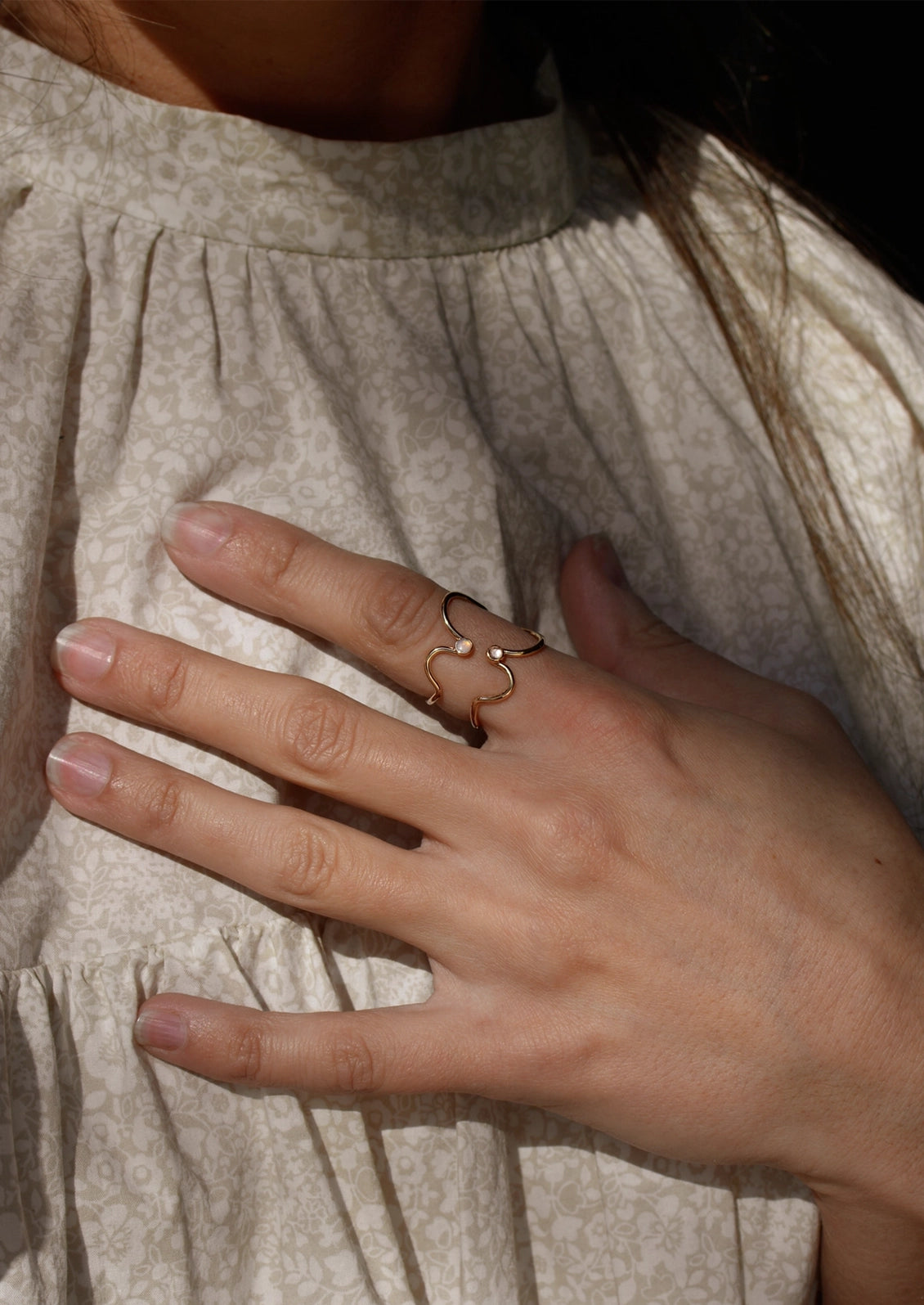 A close-up of a person's hand wearing a delicate rose gold ring with a small central bead and curved side details, resting on their cream-colored patterned dress.