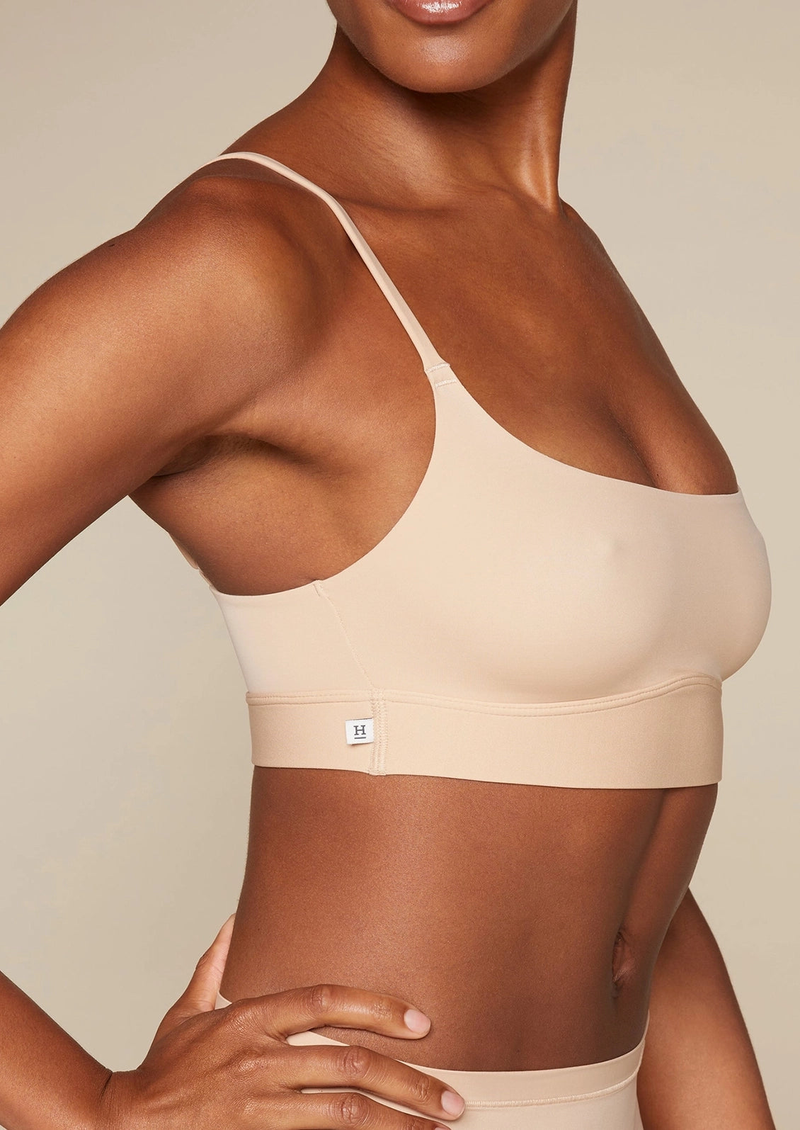 Close-up of a model wearing a light beige seamless bra with thin straps and a wide underband, shown against a neutral beige background.