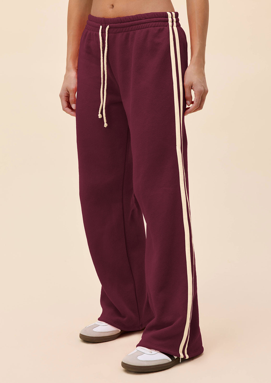 A person wearing loose, wide-leg sweatpants in deep burgundy with two cream side stripes and matching drawstrings, paired with white sneakers, against a solid light beige studio background.