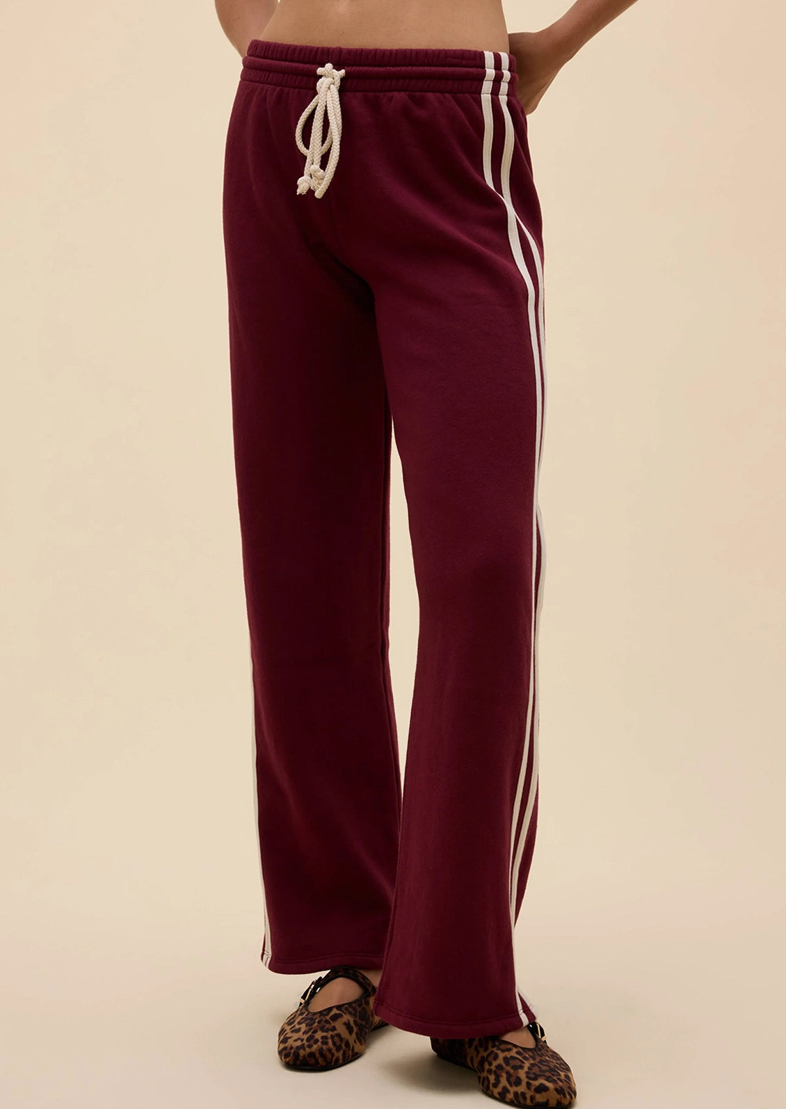 A person wearing burgundy wide-leg sweatpants with white side stripes and a white drawstring, paired with leopard-print flats, against a plain beige background.
