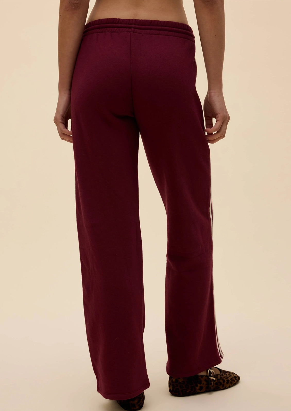 A person shown from the back wearing burgundy wide-leg sweatpants with white side stripes and leopard-print slip-on shoes, against a plain beige studio background.