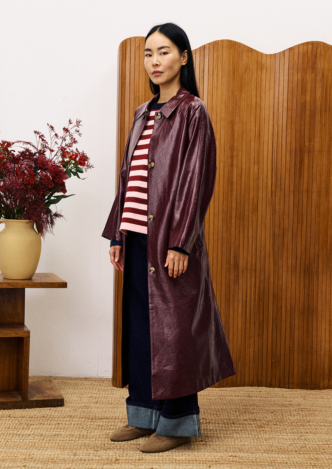 Woman wearing a long dark coat over a striped shirt, standing in front of a wooden screen with a vase of flowers on a table.