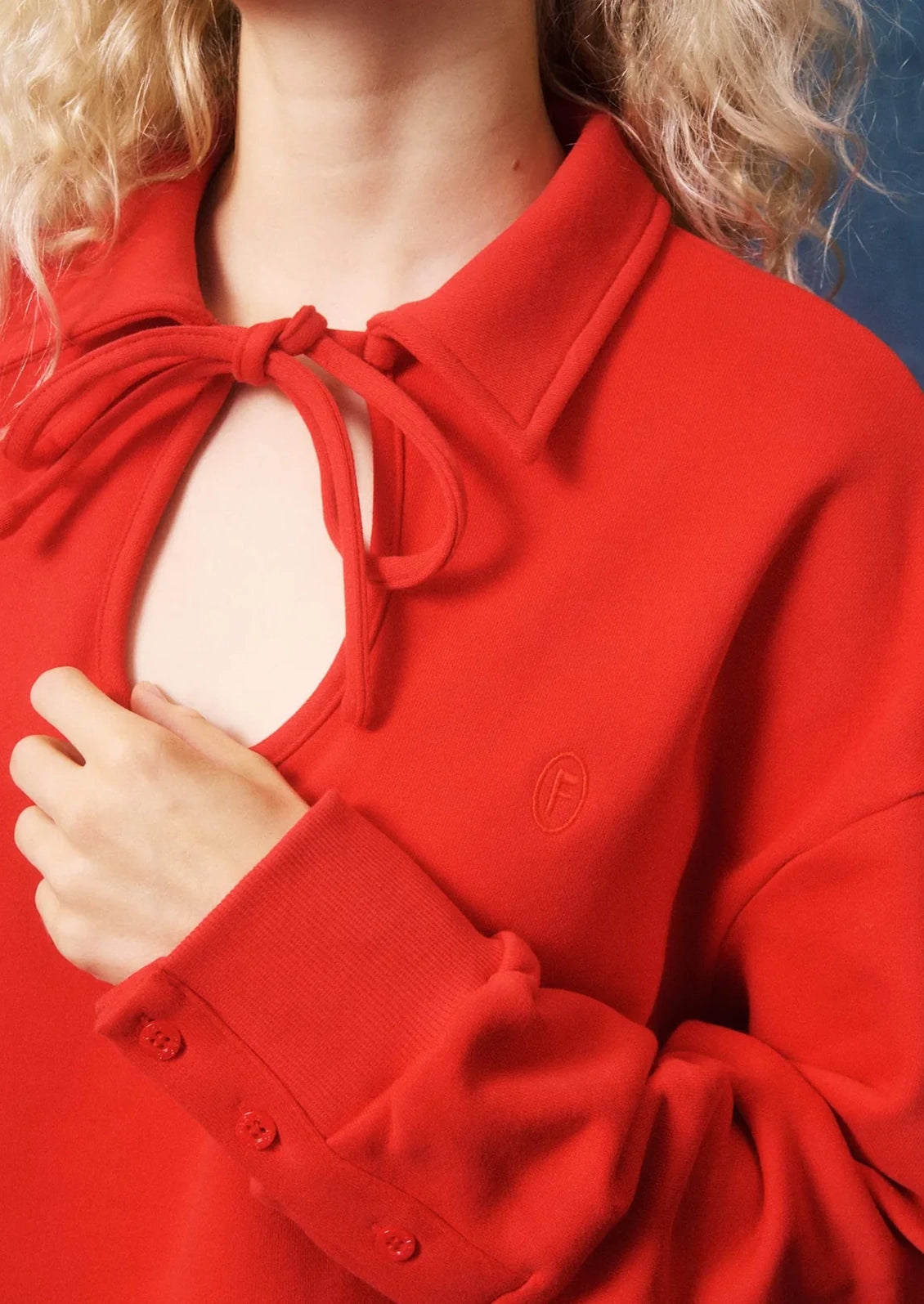 A close-up of a person wearing a bright red knit top with a collar, tie detail at the neckline, and a small tonal embroidered logo on the chest.
