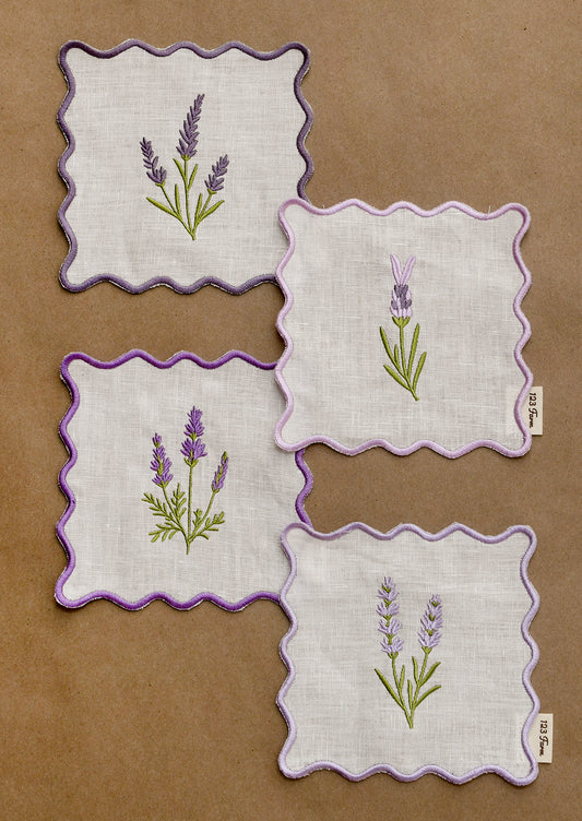 A set of four square cocktail napkins with scalloped purple edges and lavender sprig embroidery.