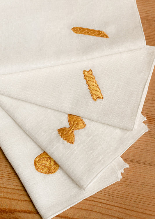 White linen napkins with embroidered pasta motifs in yellow thread, arranged on a wooden surface.