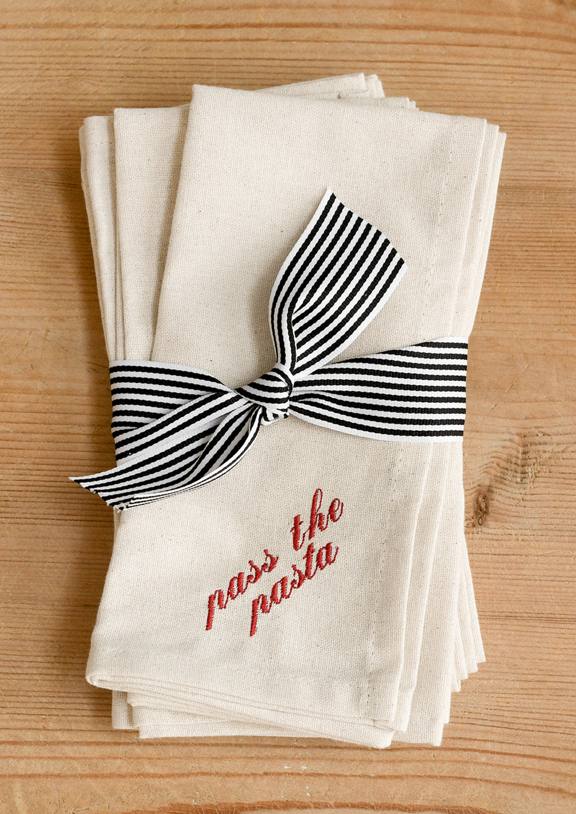 A stack of cotton napkins tied with a black and white striped ribbon, featuring the red embroidered text 'pass the pasta' on the front.