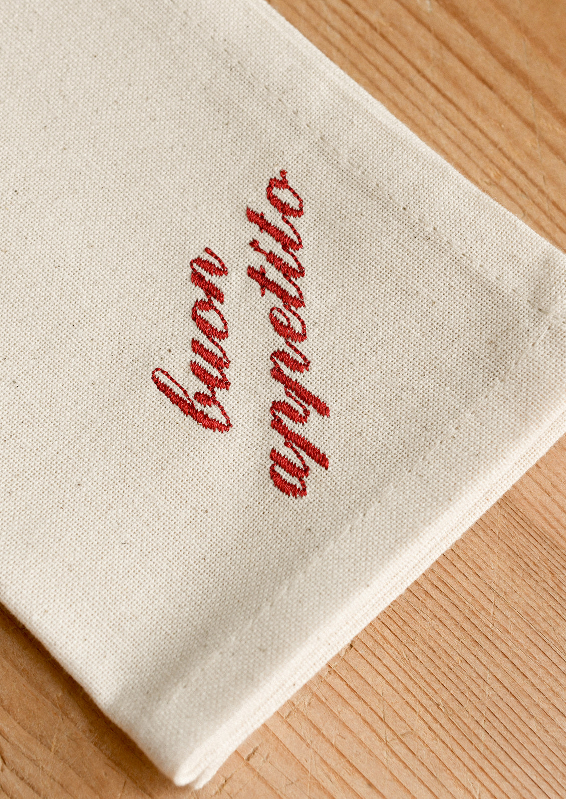 A close-up of a folded cream-colored fabric napkin with 'buon appetito' embroidered in red script, resting on a light wood surface.