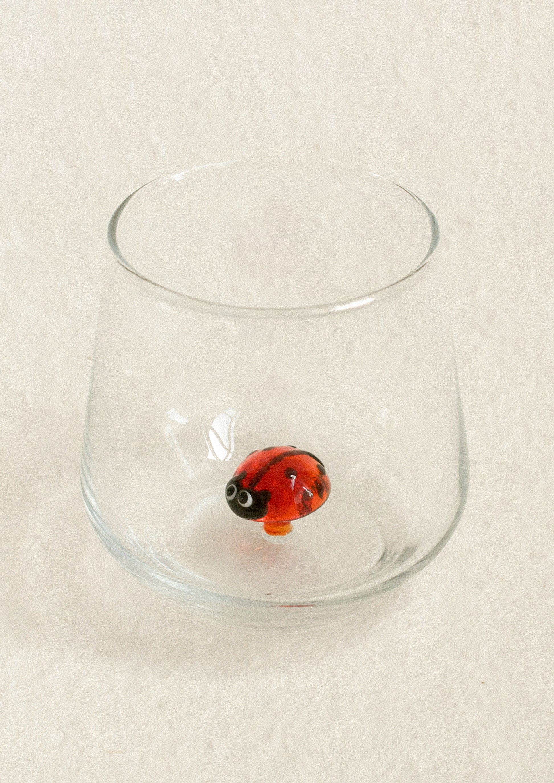 A small clear glass tumbler with a rounded base, photographed on a light beige textured background. Inside the glass sits a glossy red-and-black ladybug bead ornament.