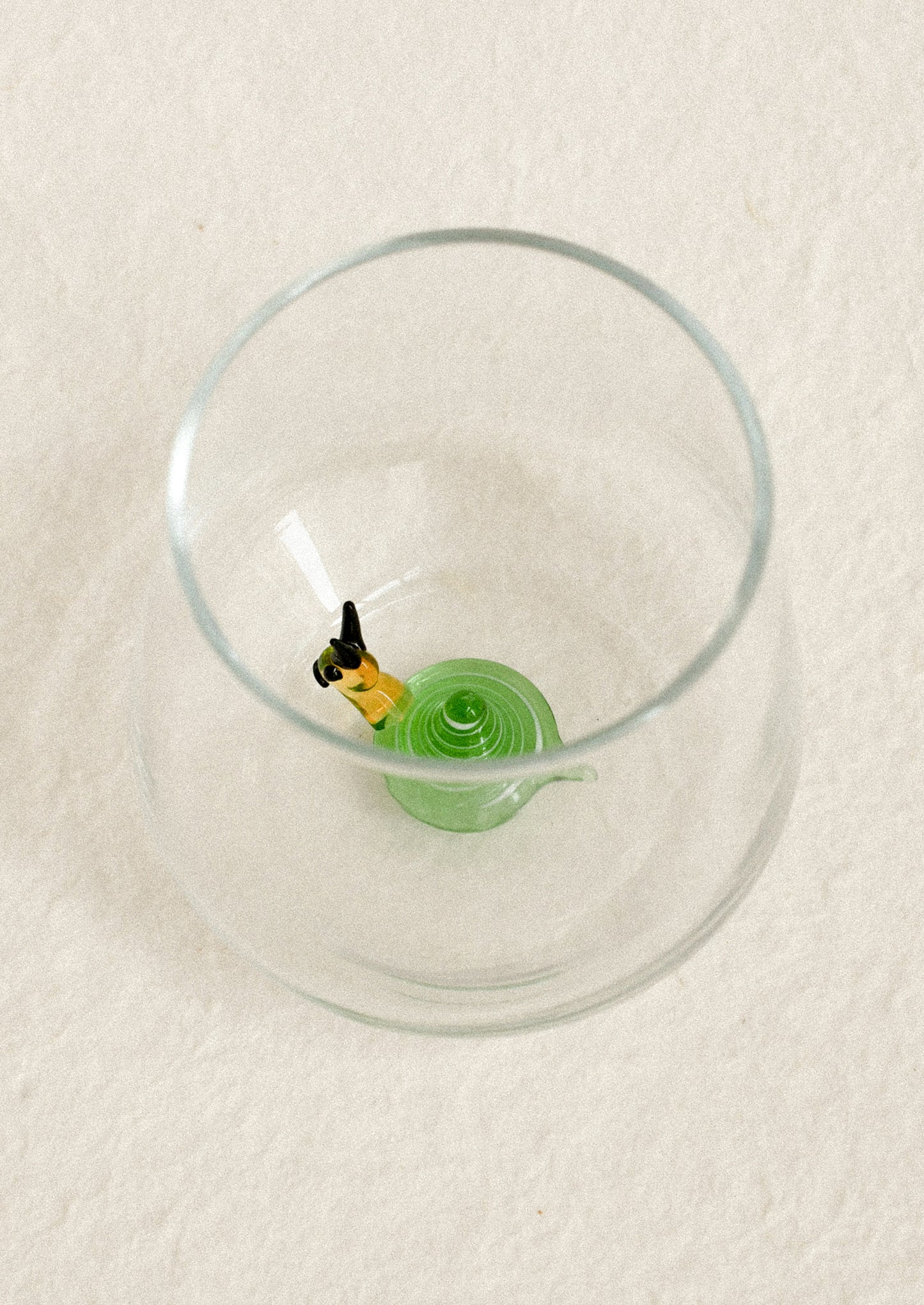 A clear glass tumbler with a small green snail detail near the bottom, photographed from above on a light textured background.