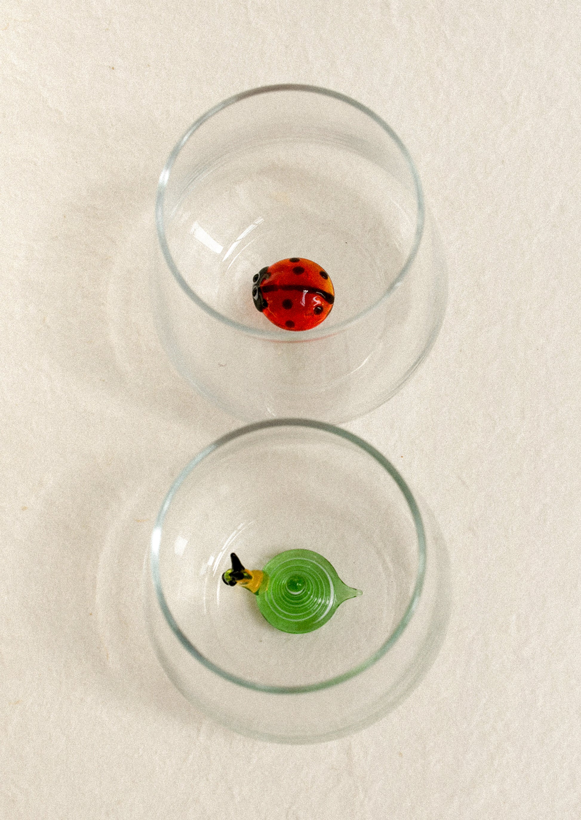 Two clear glasses, each containing a small decorative glass figure: a red ladybug in the top glass and a green snail form in the bottom style.