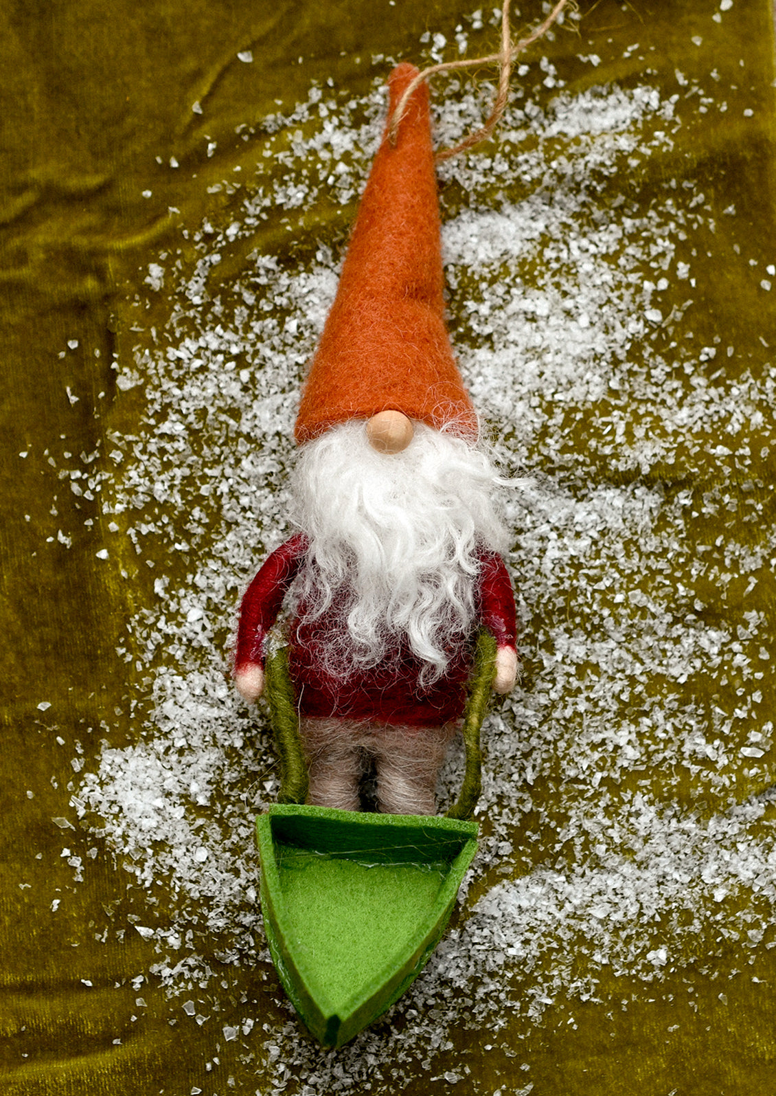 A felt Christmas gnome ornament standing in a small green boat, decorated with white faux snow on a textured olive green background.
