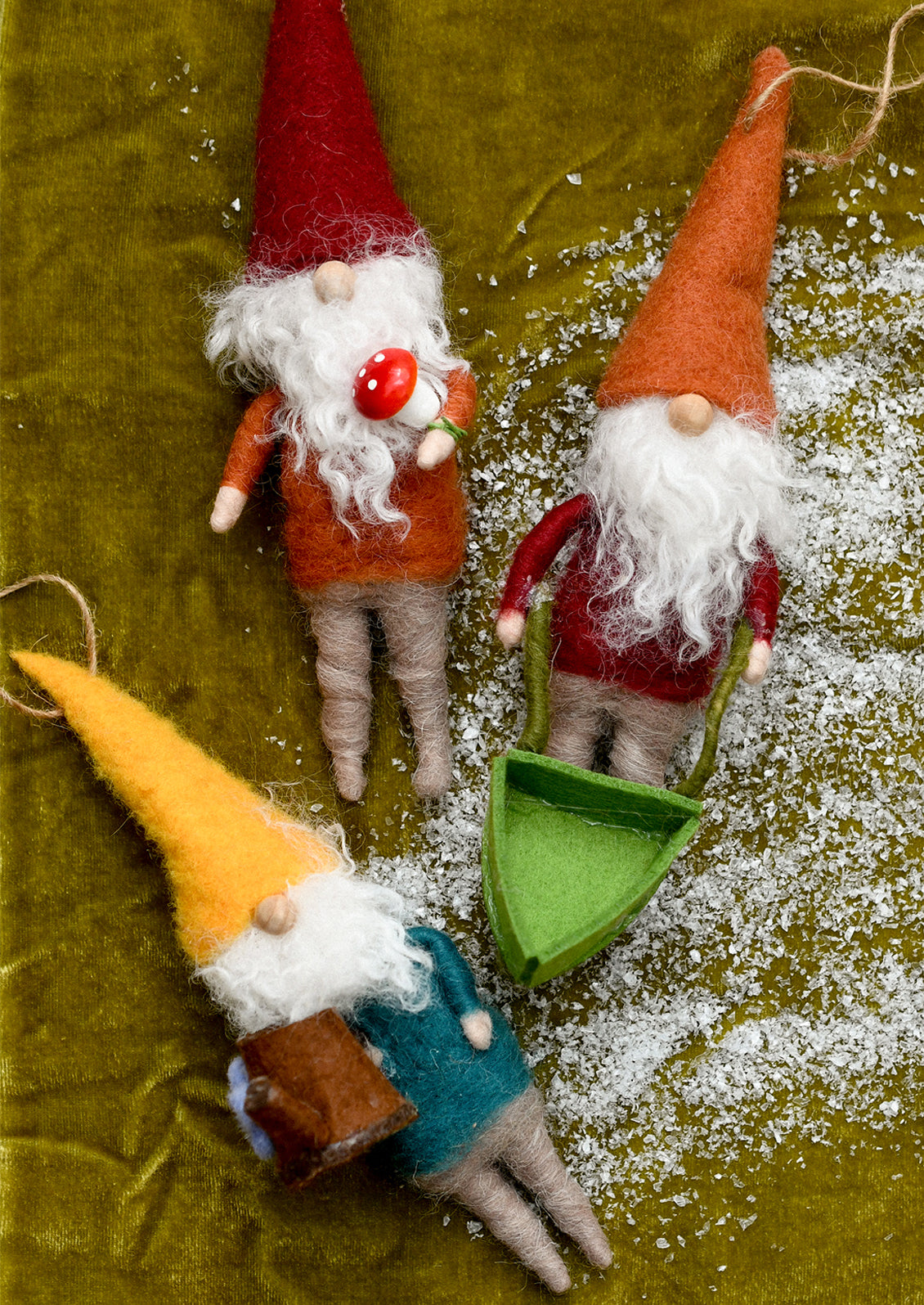 Three small felt gnome ornaments arranged on an olive green fabric background with white faux snow. One gnome wears a red hat and orange outfit and holds a red ornament; another wears an orange hat and red outfit and sits in a green sled; the third wears a yellow hat and teal outfit and holds a brown object.