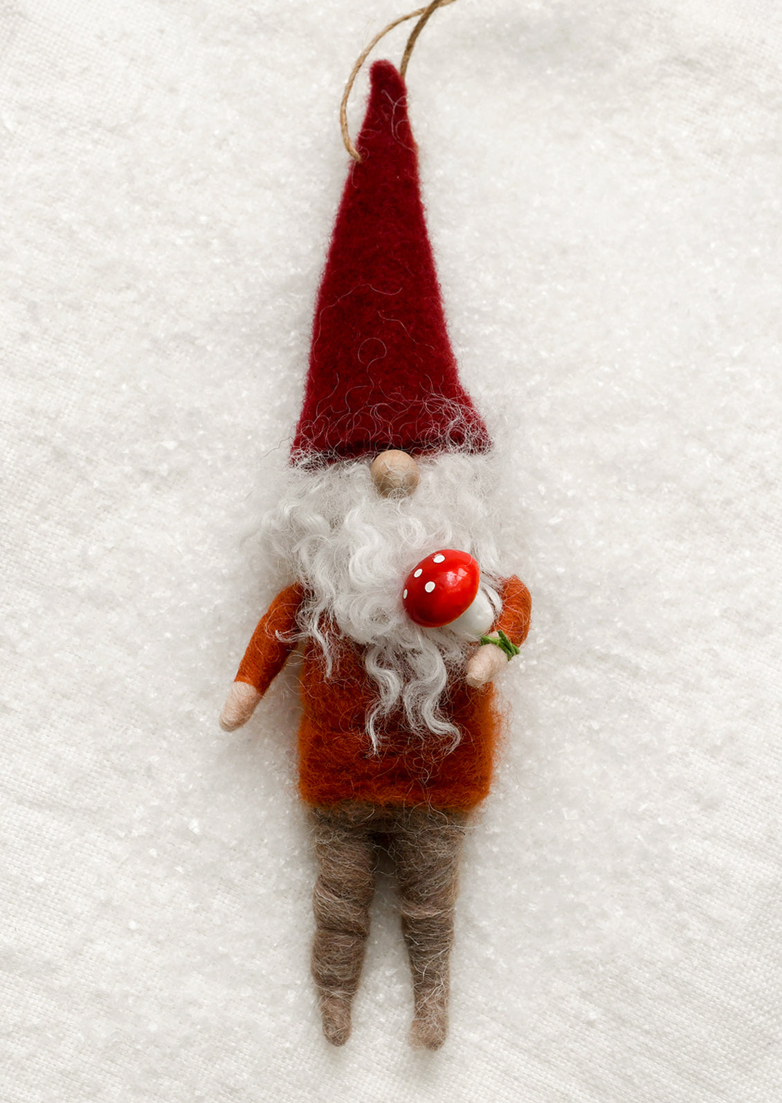 A handcrafted felted wool gnome ornament hanging against a textured white fabric background. The gnome features a tall red hat, a long white beard, an orange-brown shirt, brown felted legs, and holds a small red-and-white mushroom.