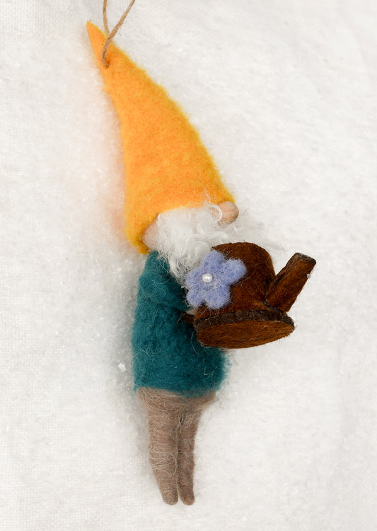 A handcrafted felted wool gnome ornament lying on a textured white fabric background. The gnome features a bright orange-yellow pointed hat, a white beard, a teal-green sweater, brown pants, and holds a small purple flower with a silver bead in its hand.