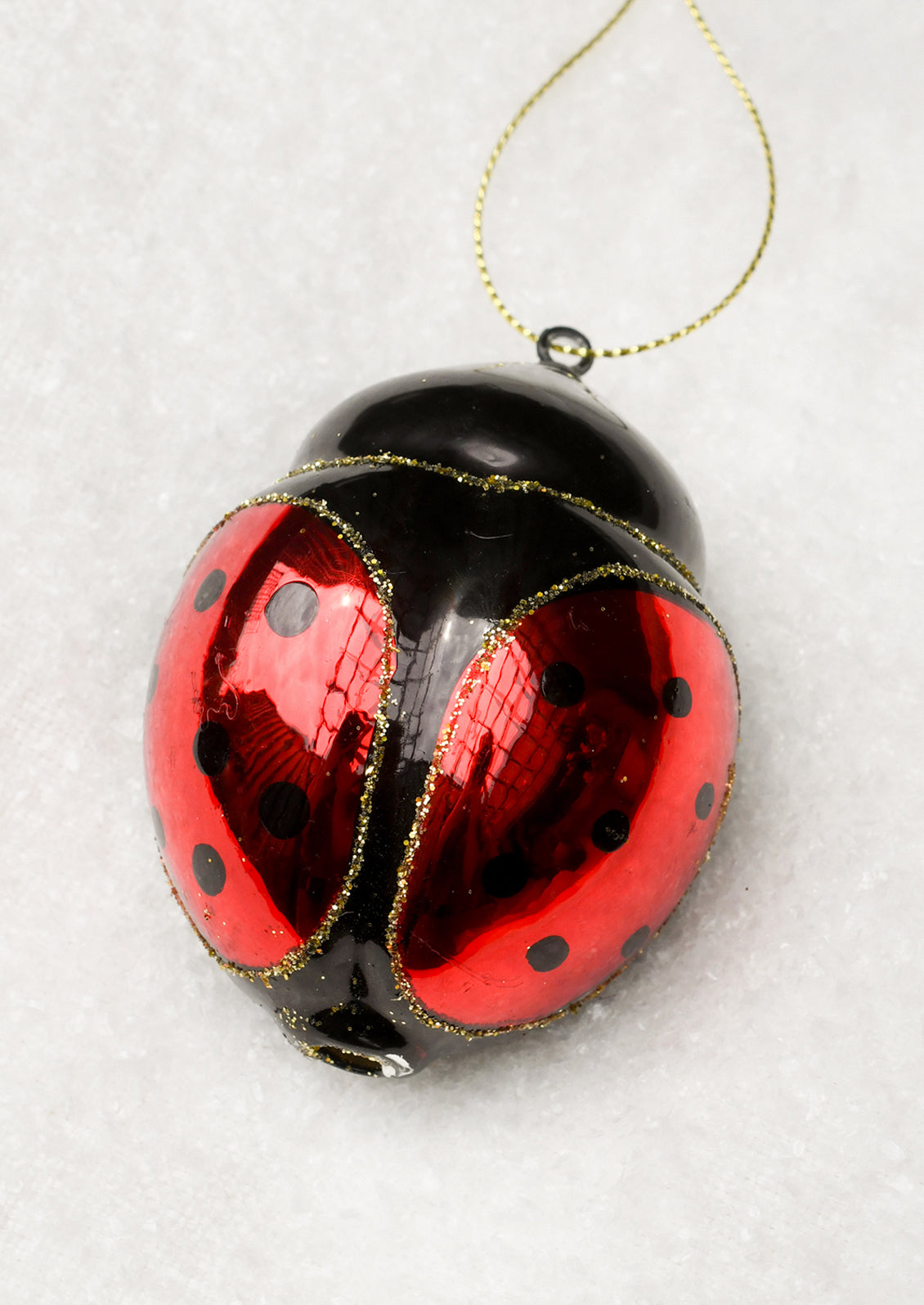 A glossy ladybug-shaped Christmas ornament with red and black sections, gold glitter edging, and a gold hanging string, photographed on a white textured surface.