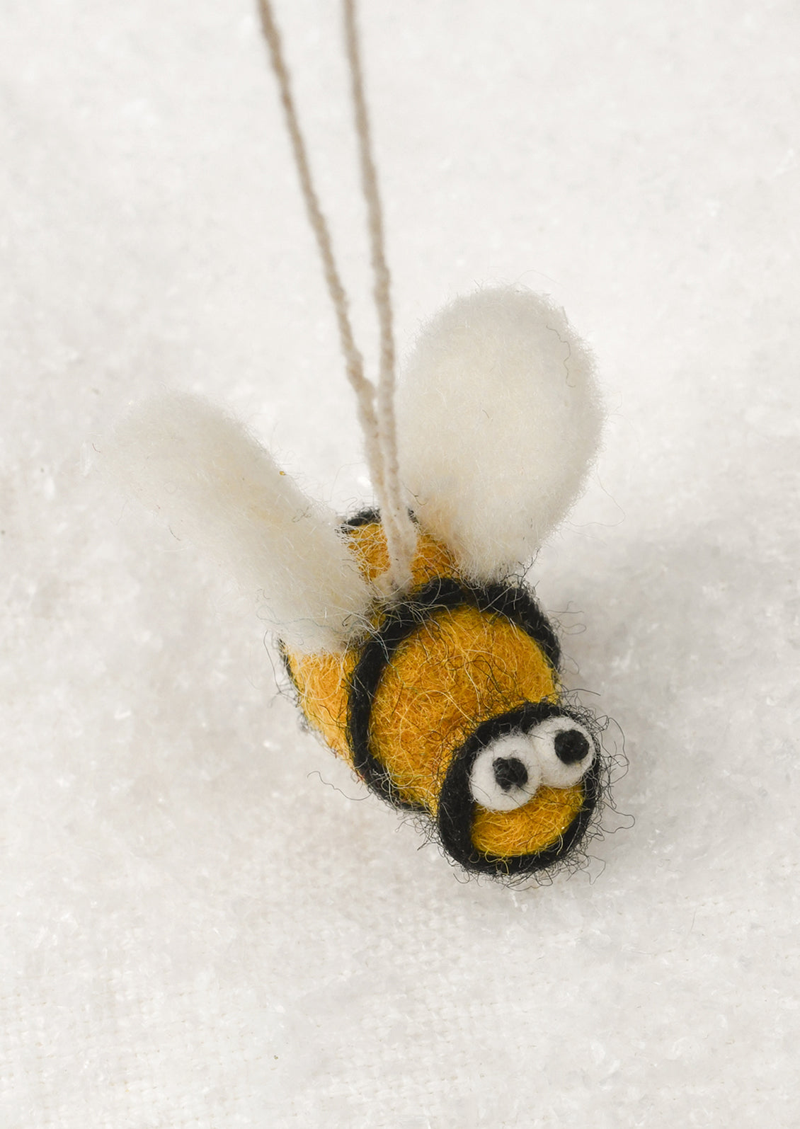 A small felted wool bumblebee ornament with white wings, yellow and black stripes, and googly eyes, hanging against a soft white textured background.