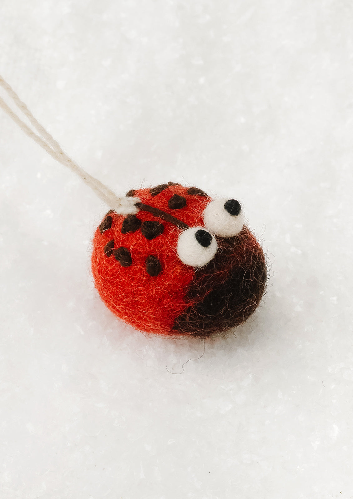 A small felt ladybug ornament with red and black felted wool body, white googly eyes, and a white hanging string, photographed on a white textured surface.