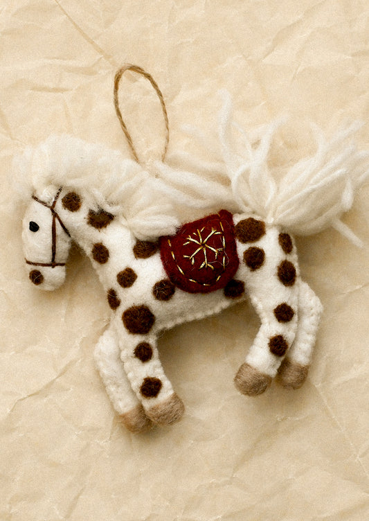 A handmade felt Christmas ornament shaped like a white horse with brown spots, a red saddle with a gold snowflake, and white yarn mane and tail, hanging from a twine loop.
