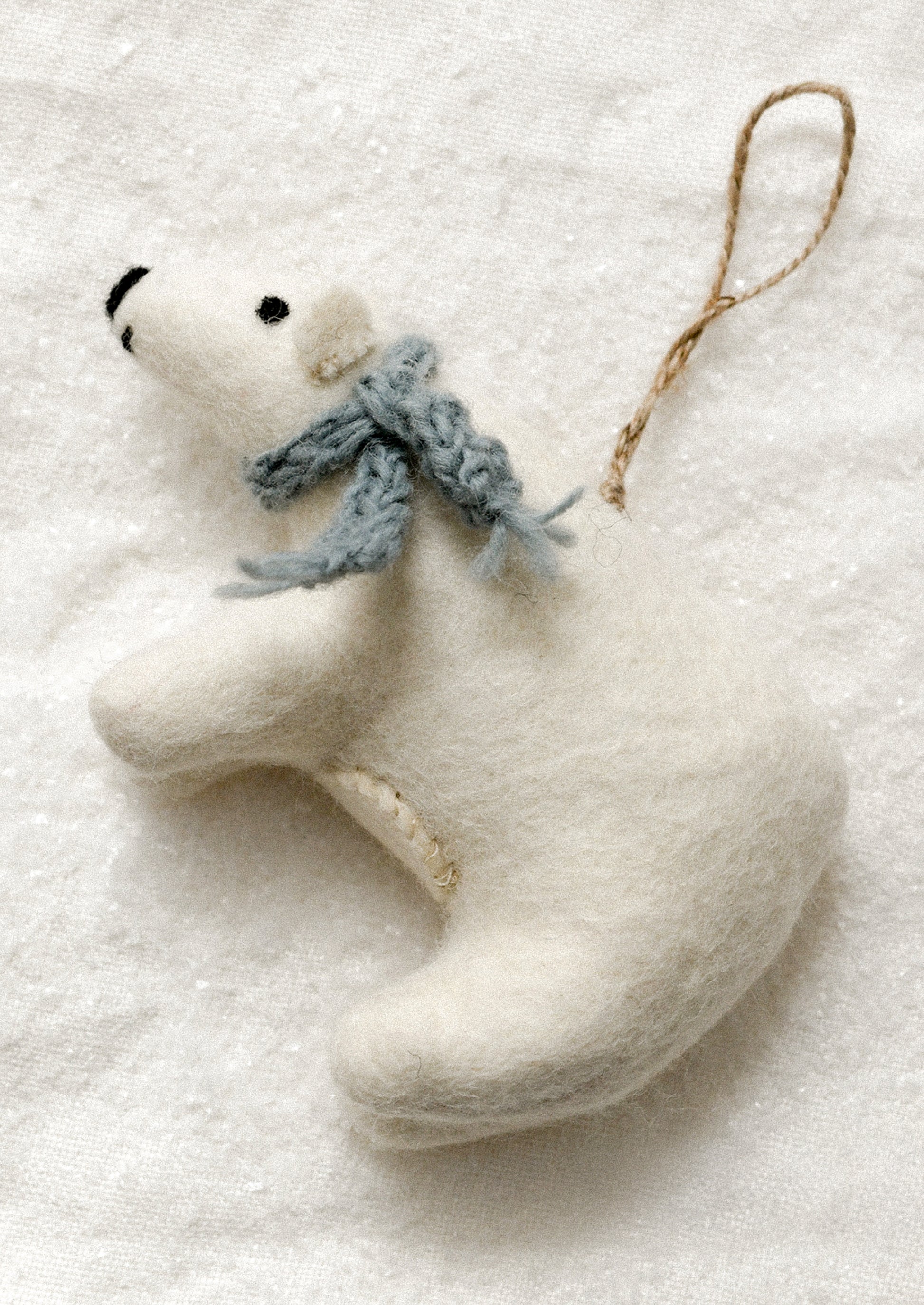 A white felt polar bear Christmas ornament with a knitted blue-gray scarf and a brown jute hanging loop, resting on a textured white background.