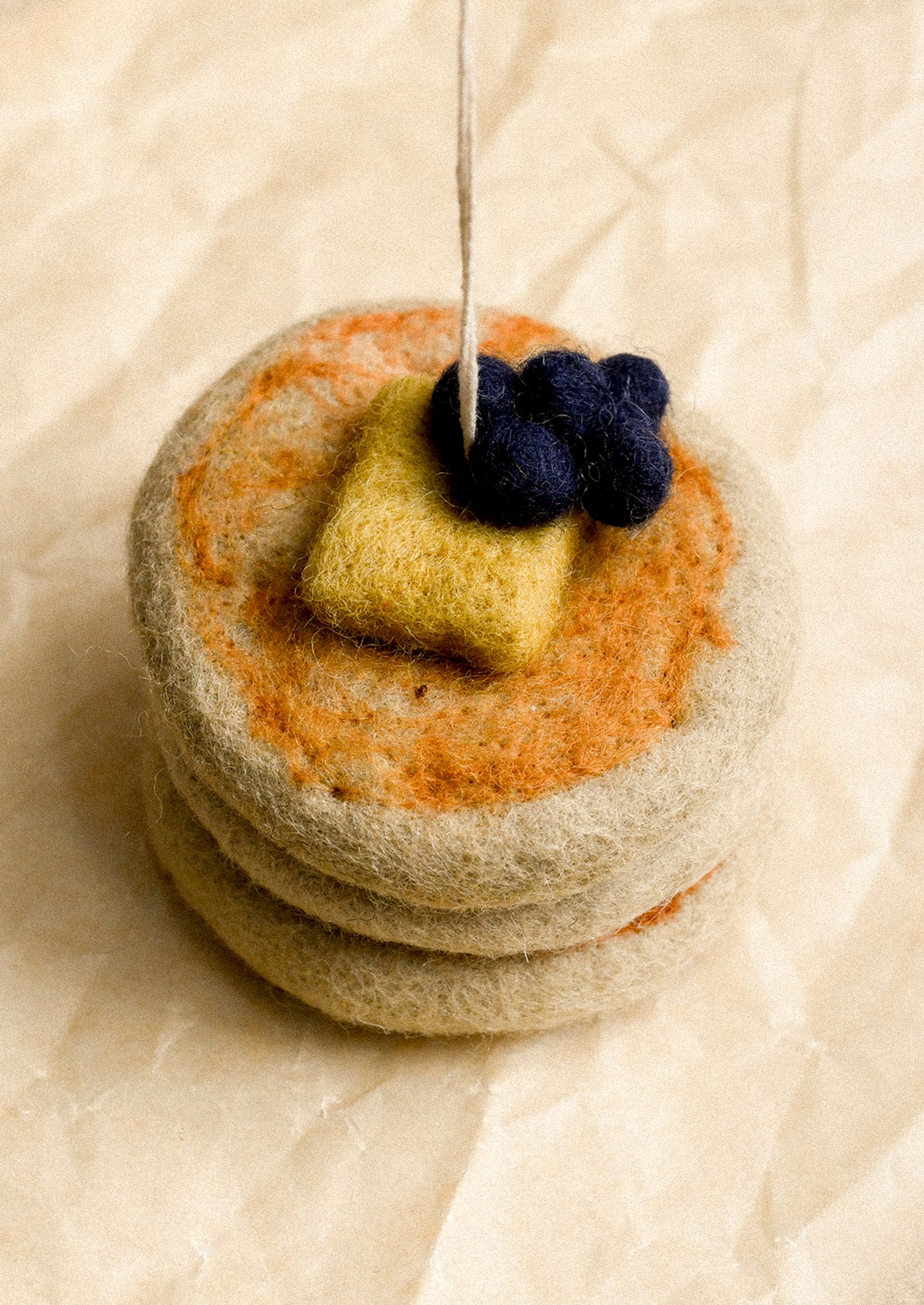 A felted wool miniature of a stack of pancakes topped with butter and blueberries, resting on a textured beige paper background.