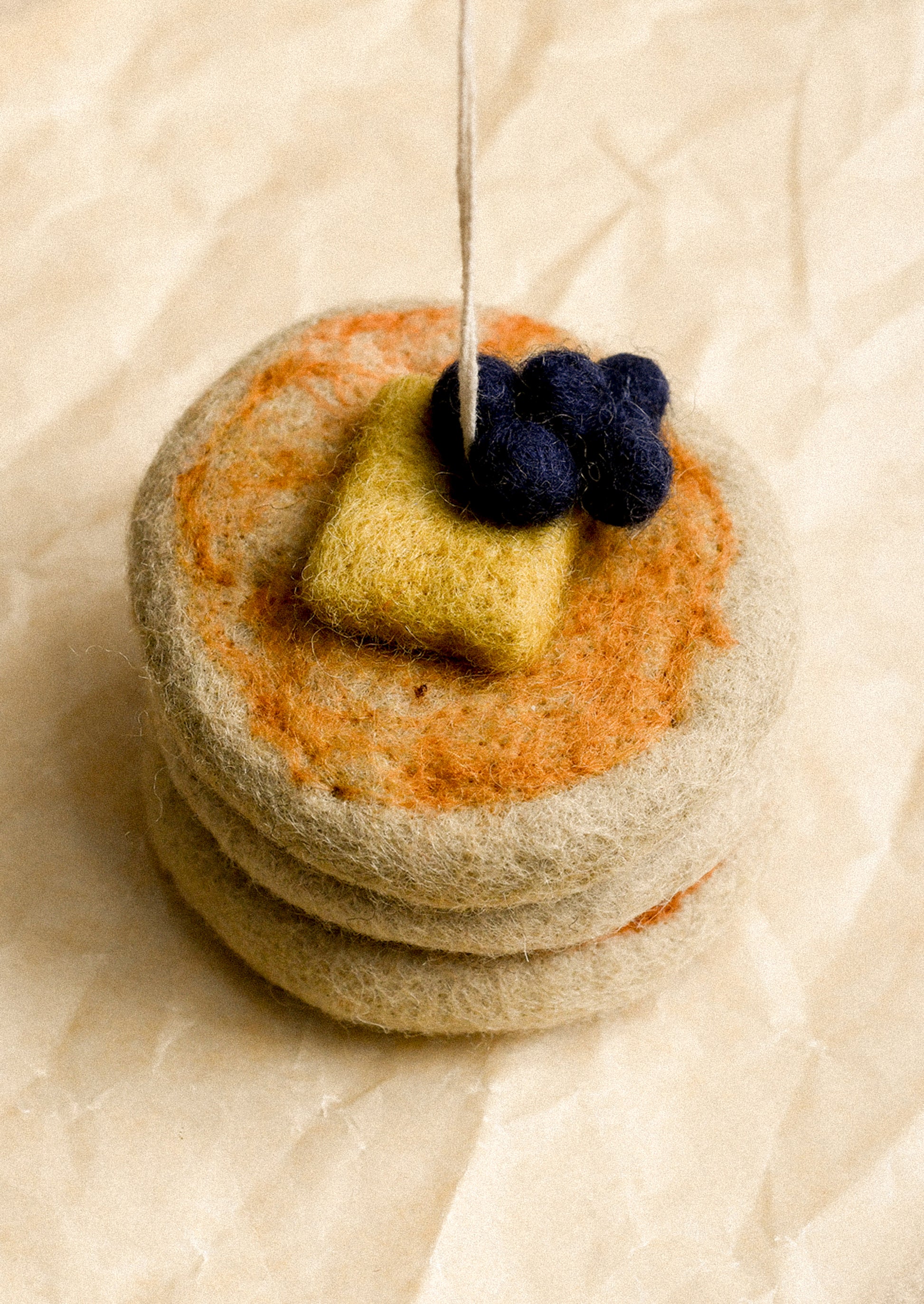 A felted wool miniature of a stack of pancakes topped with butter and blueberries, resting on a textured beige paper background.